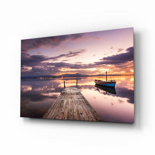 Wooden jetty extending toward calm water at sunset with a moored blue and black boat, mountains visible across the bay under purple and orange sky, displayed as glass wall art.