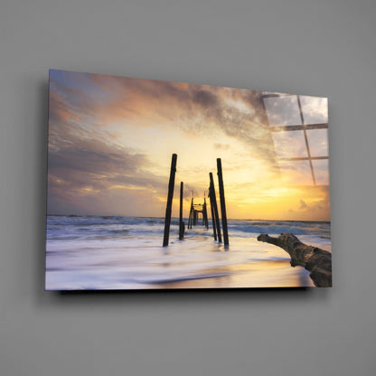 Sunset beach scene with wooden pier pilings and driftwood printed on glass wall art mounted on gray wall.