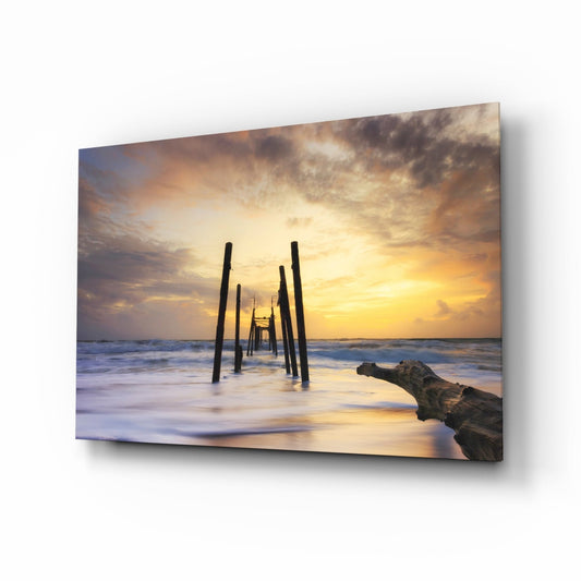 Sunset beach scene with silhouetted wooden pier pilings, golden sky, waves, and driftwood on glass wall art.