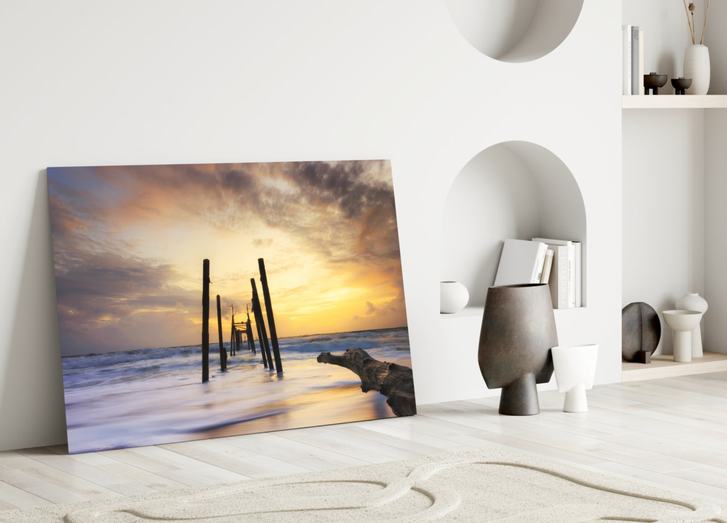 Glass wall art leaning against white wall displays sunset beach scene with wooden pier pilings and golden sky.