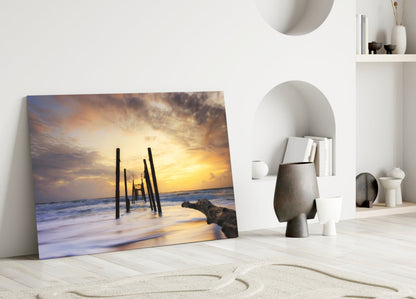 Glass wall art leaning against white wall displays sunset beach scene with wooden pier pilings and golden sky.