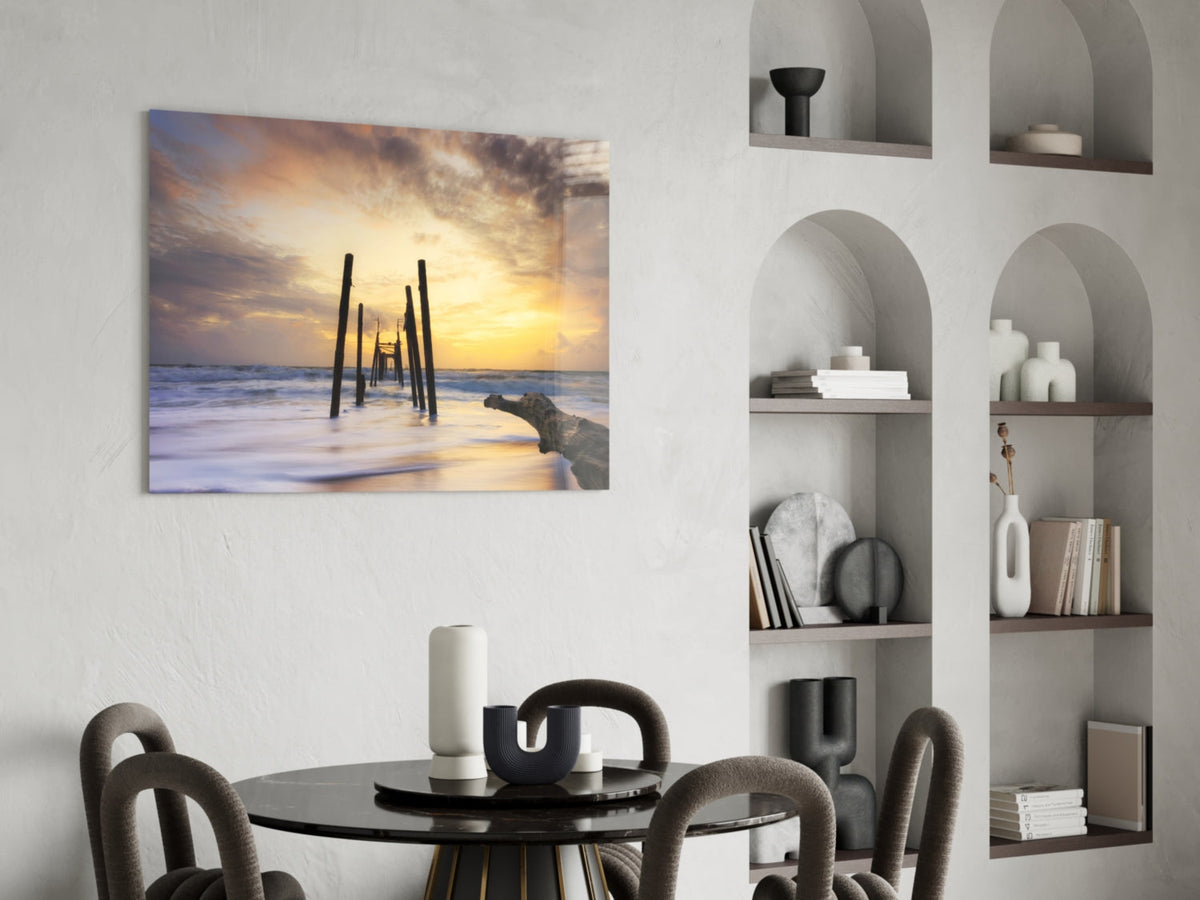 Canvas print of a sunset pier extending into the ocean with golden sky and blue waves, displayed above a black dining table in a modern interior.