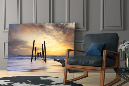 Destroyed Dock Glass Wall Art leaning against wall beside wooden chair, displaying sunset beach pier scene with golden sky and silhouetted pilings.