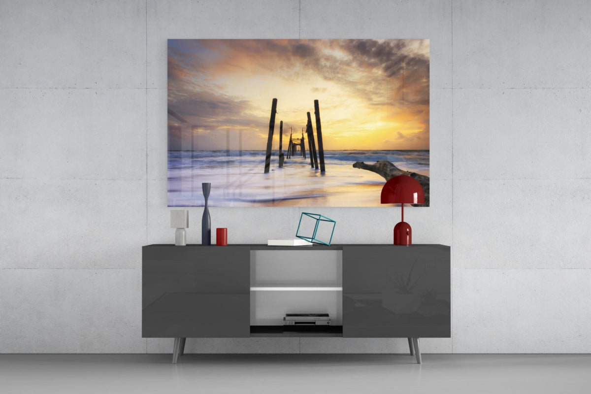 Glass wall art depicting a sunset pier with wooden posts and beach scene, mounted above gray media console with decorative accessories.