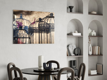 Birdcage glass wall art hanging above a round black dining table with beige chairs, showing decorative bird cages with purple flowers and warm sepia tones.