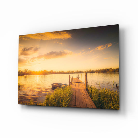 Glass wall art panel displayed at an angle showing a wooden dock extending into a golden-lit lake at sunset with a rowboat nearby.