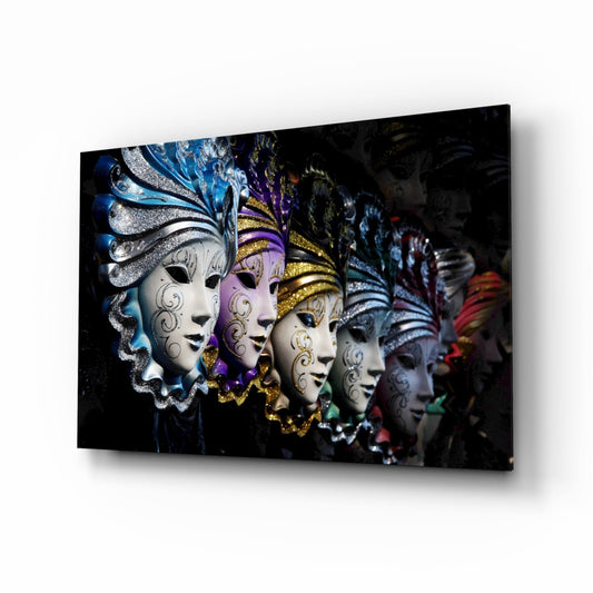Angled perspective of glass wall art displaying five ornate Venetian carnival masks with white faces, decorated with blue, purple, and gold embellishments against a dark background.