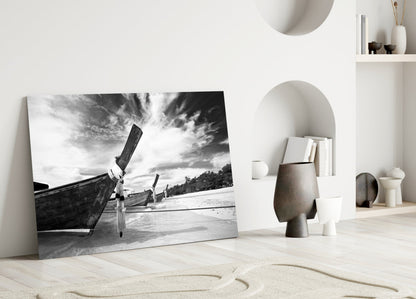 Black and white glass wall art leaning against white wall displaying traditional boats on beach with dramatic sky.