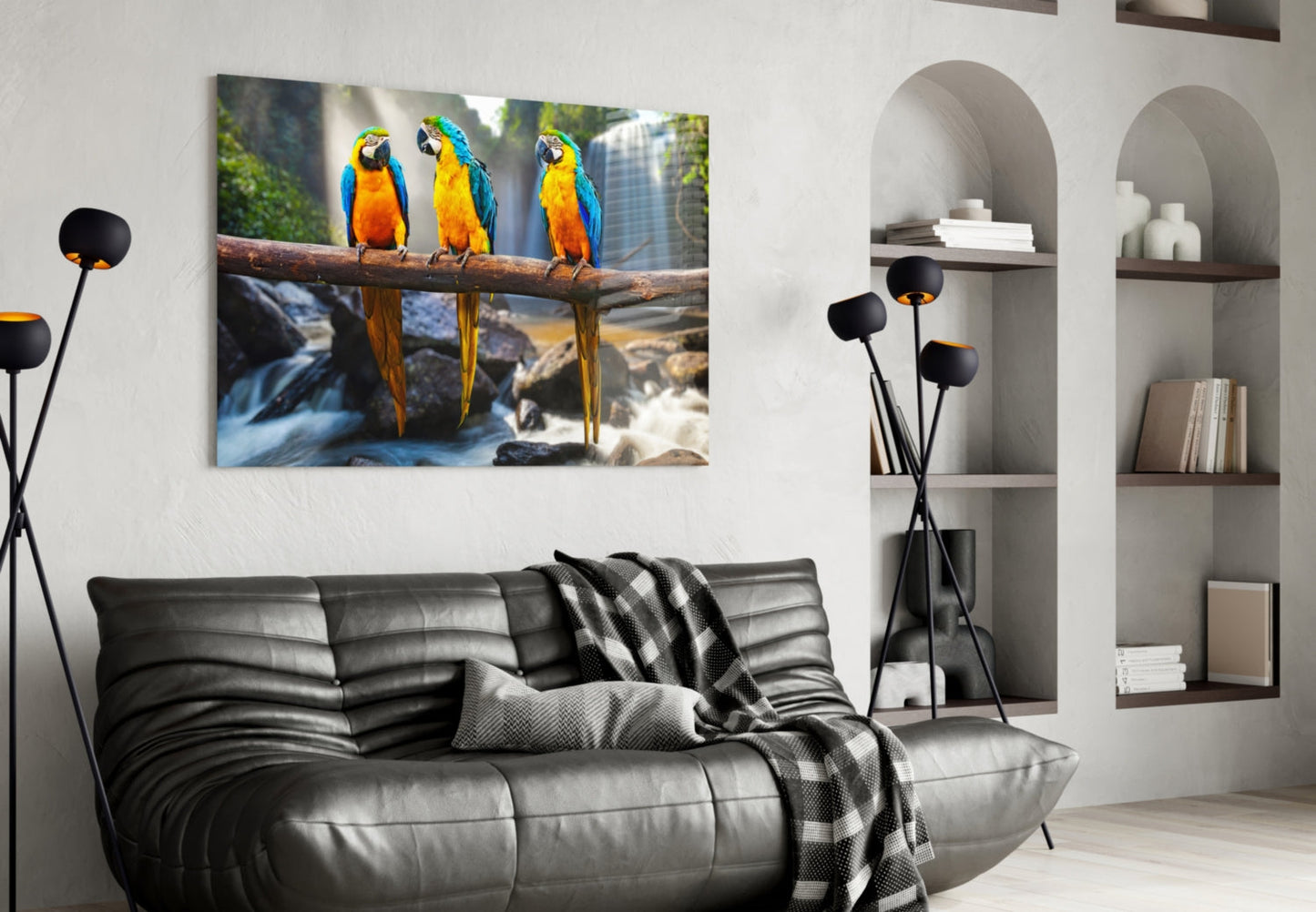 Glass wall art depicting three blue and gold macaws perched on a branch above a waterfall, displayed above a black leather sofa in a modern living room.