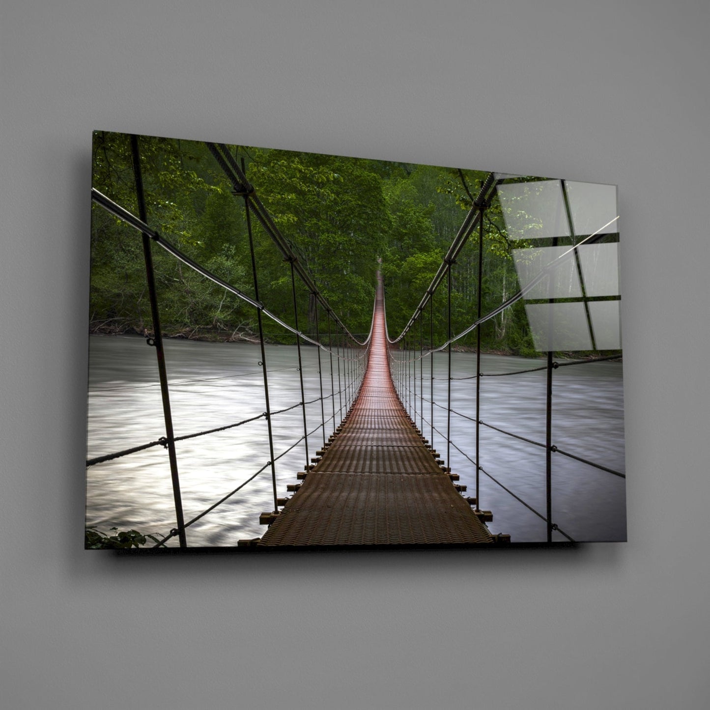 Suspension bridge glass wall art displayed on gray wall, showing perspective view down wooden walkway spanning river with green forest canopy.