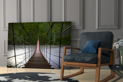 Suspension bridge glass wall art displayed in modern living room beside wooden chair with blue cushion, showing forest canopy and river perspective.