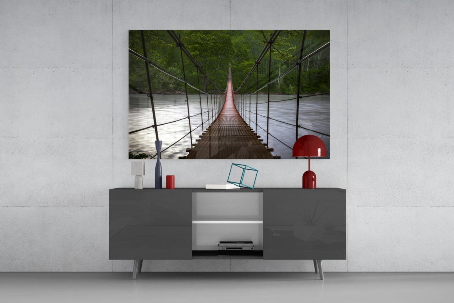 Suspension bridge glass wall art mounted above gray media console displaying long perspective view of red pedestrian bridge over river with green forest.