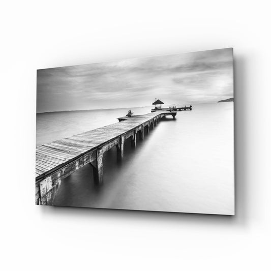 Black and white long exposure photograph of a weathered wooden pier extending into misty water with a small pavilion structure at the end, displayed as a glass wall art print.
