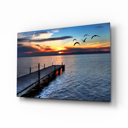 Wooden dock extending into calm water at sunset with orange sky, flying birds, and glass wall art mounted at angle.
