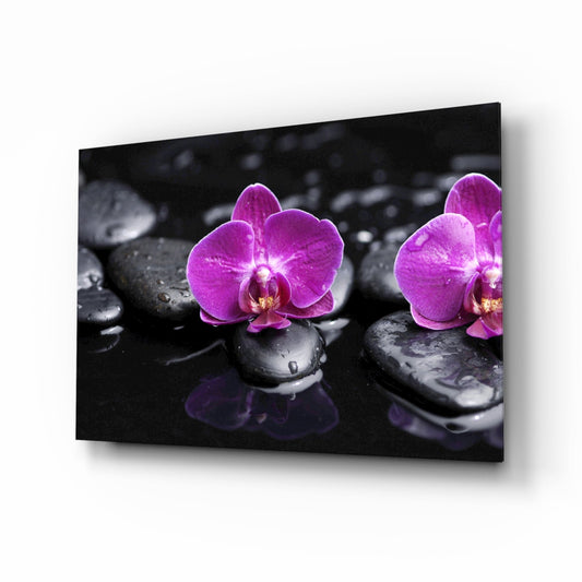 Glass wall art displaying two magenta orchids on wet black zen stones against dark background, angled perspective view.