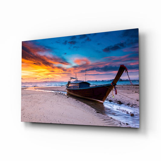 Wooden longtail boat anchored on sandy beach at sunset with vibrant orange and blue sky reflected on glass wall art panel.