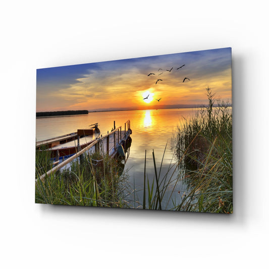 A wooden pier extends into a calm lake at golden sunset with reeds in foreground and birds flying overhead, displayed as glass wall art.