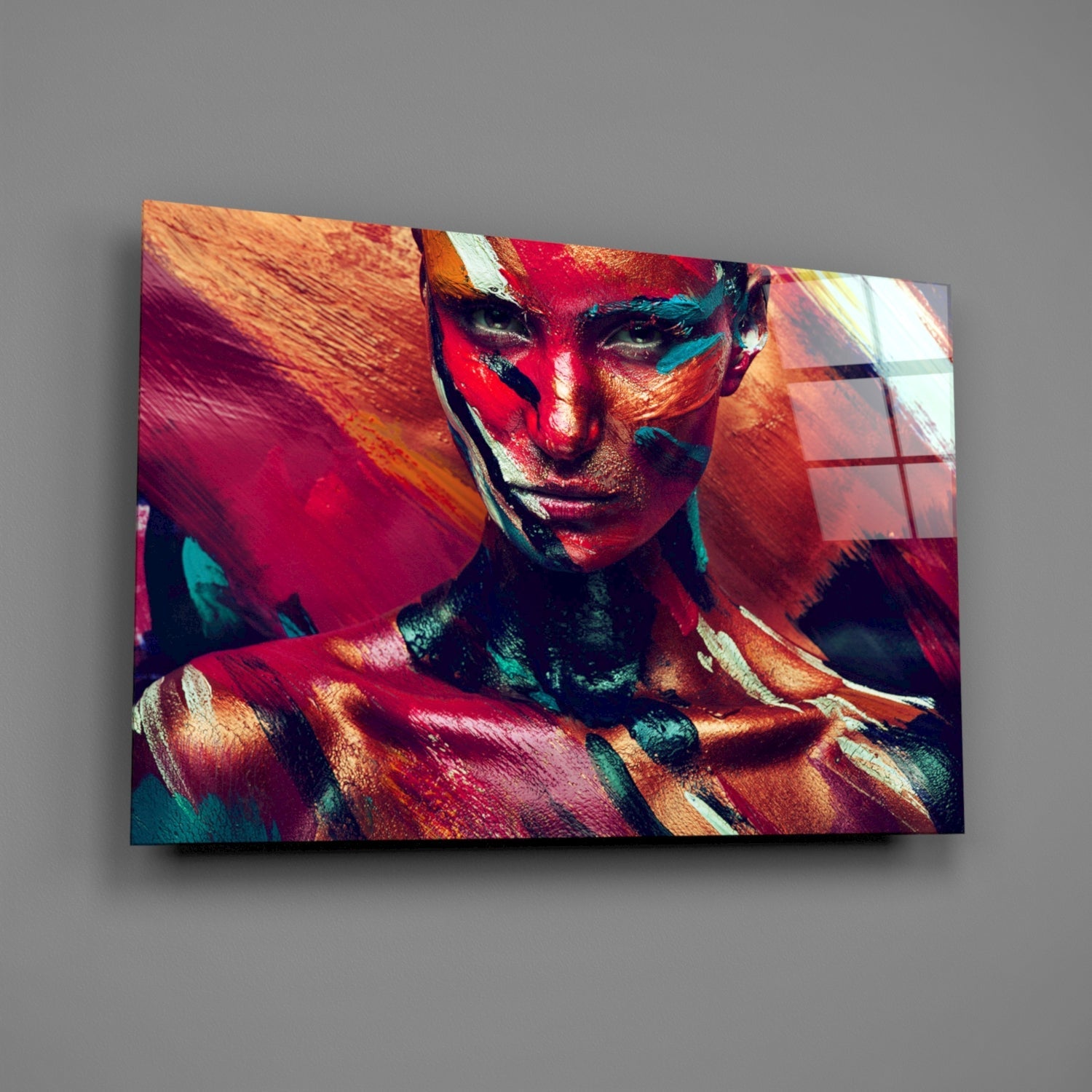 Colorful glass wall art displaying a woman's face painted in vibrant red, orange, teal, and yellow pigments, mounted at an angle on a gray wall.