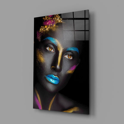Glass wall art displaying a woman's face with black skin, golden and blue streaks, magenta accents, and bright blue lips, mounted at an angle against gray background.