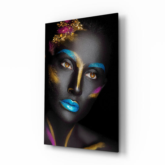 Woman's face with dark skin painted in blue, gold, and magenta streaks, golden eyes, blue lips, mounted on glass panel against white background.