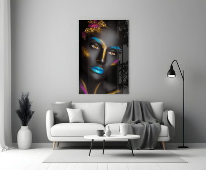 Glass wall art depicting a woman's face with dark skin, blue and gold paint, striking amber eyes, displayed above a white sofa in a modern living room.