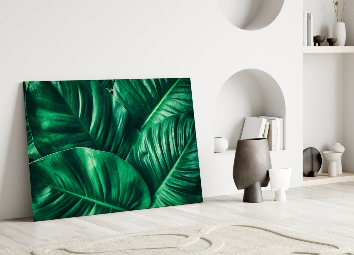 Green tropical leaf glass wall art leaning against white wall in minimalist interior with decorative objects and shelving.