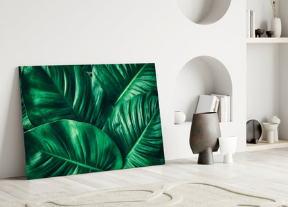 Green tropical leaf glass wall art leaning against white wall in minimalist interior with decorative objects and shelving.