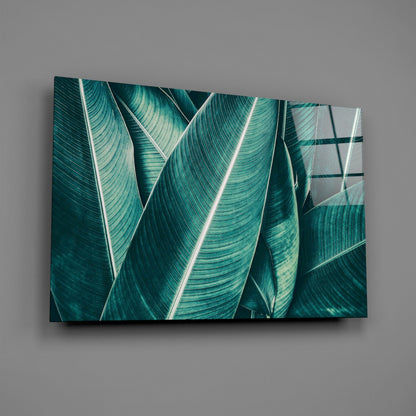 Glass wall art displaying overlapping teal and emerald tropical leaves with white veins against a gray background.