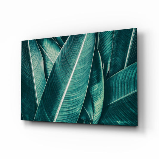 Angled perspective view of glass wall art displaying layered teal and dark green tropical leaves with white veins against white background.