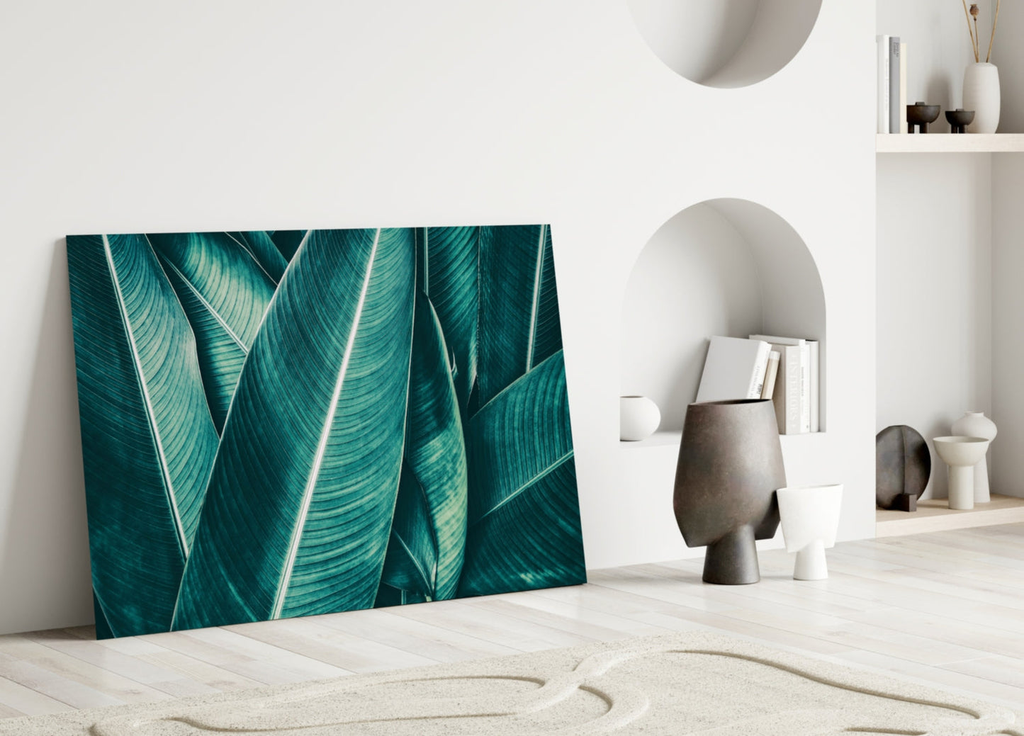 Green tropical leaf glass wall art leaning against white wall in minimalist room setting with decorative objects.