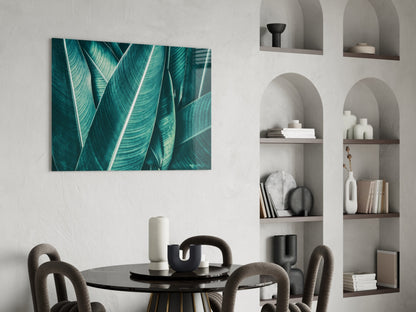 Teal tropical leaf glass wall art displayed above a black dining table in a modern interior with white built-in shelving.