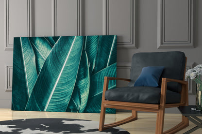 Green tropical leaf glass wall art displayed in modern living room with wooden chair and grey paneled wall.