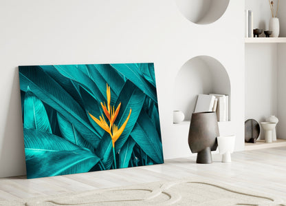Tropical Leaf 3 glass wall art leaning against white wall, displaying turquoise leaves with bright yellow-orange bird of paradise flower.
