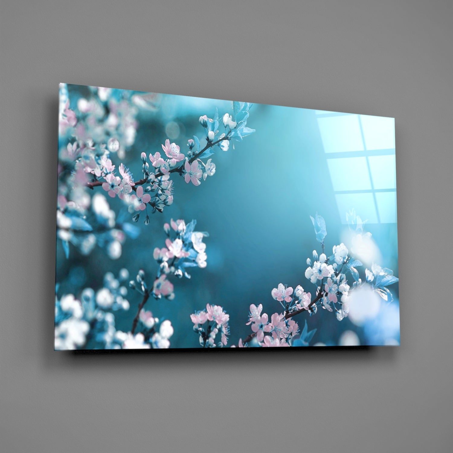 Glass wall art mounted on gray wall displaying pink and white spring blossoms against turquoise sky with window reflection.