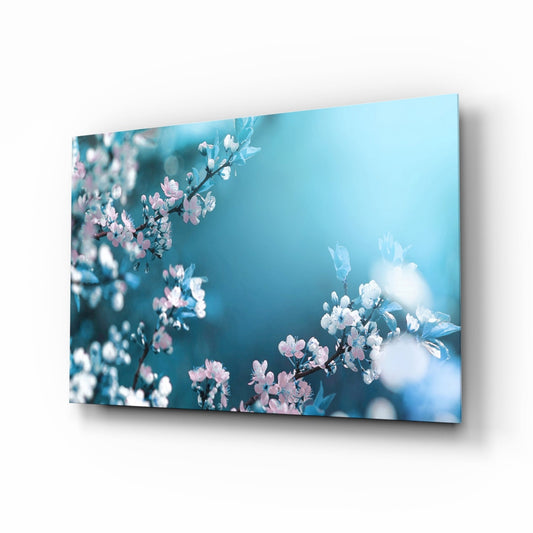 Glass wall art panel displayed at an angle showing pink and white spring blossoms against a soft blue background.