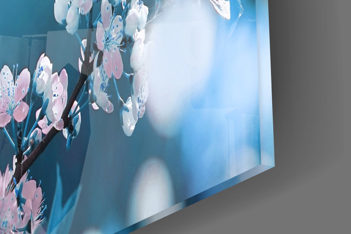 Glass wall art panel displaying pink and white cherry blossoms on branches against a blue gradient background, angled perspective view.