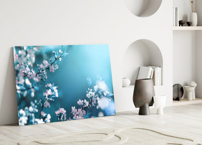 Glass wall art leaning against white wall displays pink and white flowers against turquoise blue background.