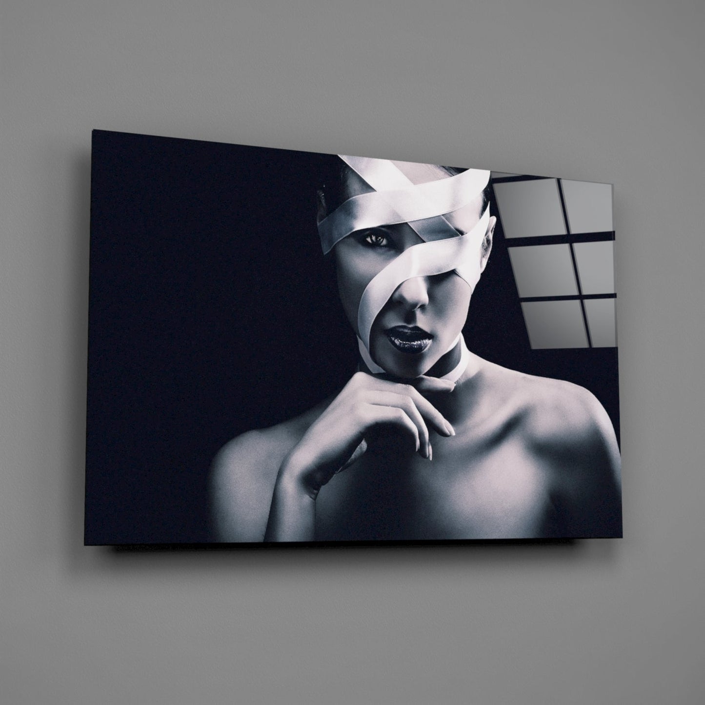 Monochromatic portrait of a woman with white ribbon-like strips across her face on black glass panel, mounted on gray wall.