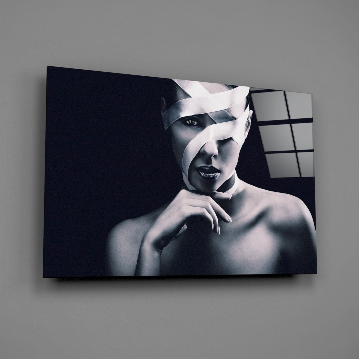 Monochromatic portrait of a woman with white ribbon-like strips across her face on black glass panel, mounted on gray wall.