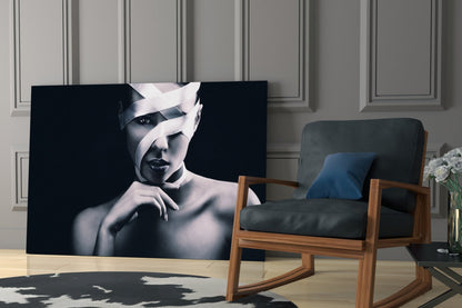 Black and white portrait of woman with white fabric wrapped around head, displayed as glass wall art in modern living room with wooden chair.