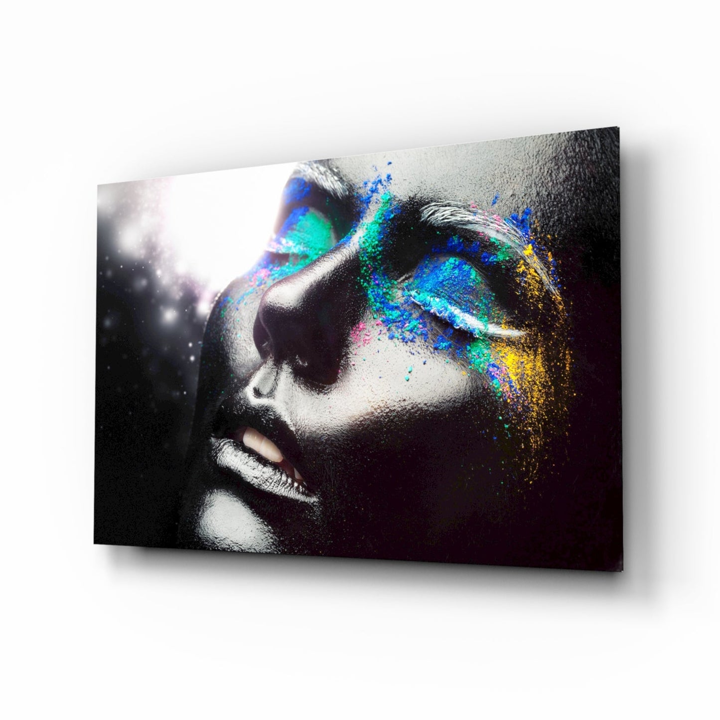 Profile view of female face in black and white with vibrant blue, cyan, yellow, and magenta powder dispersing across right side on glass panel.