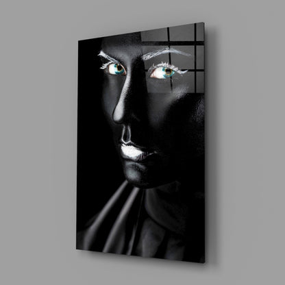 Black and white glass portrait with striking turquoise eyes, illuminated face against dark background, mounted on gray wall.