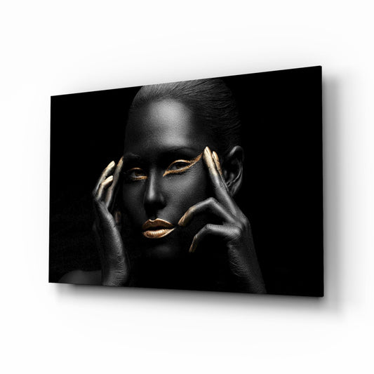 Woman with black body paint and gold lips resting chin on hands against dark background, displayed as angled glass wall art.