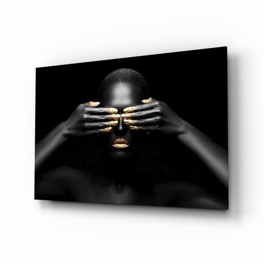 Black and gold glass wall art showing a woman's face obscured by her hands with gold-painted fingernails and lips against dark background.