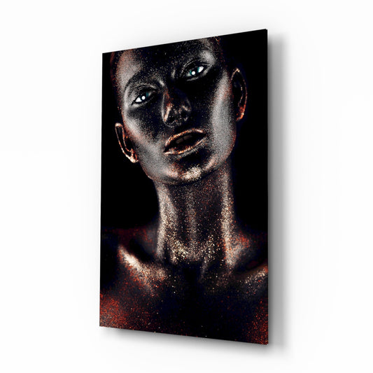 Angled view of glass wall art depicting a woman's face with glowing white eyes, copper and gold glitter finish against dark background, mounted on white wall.
