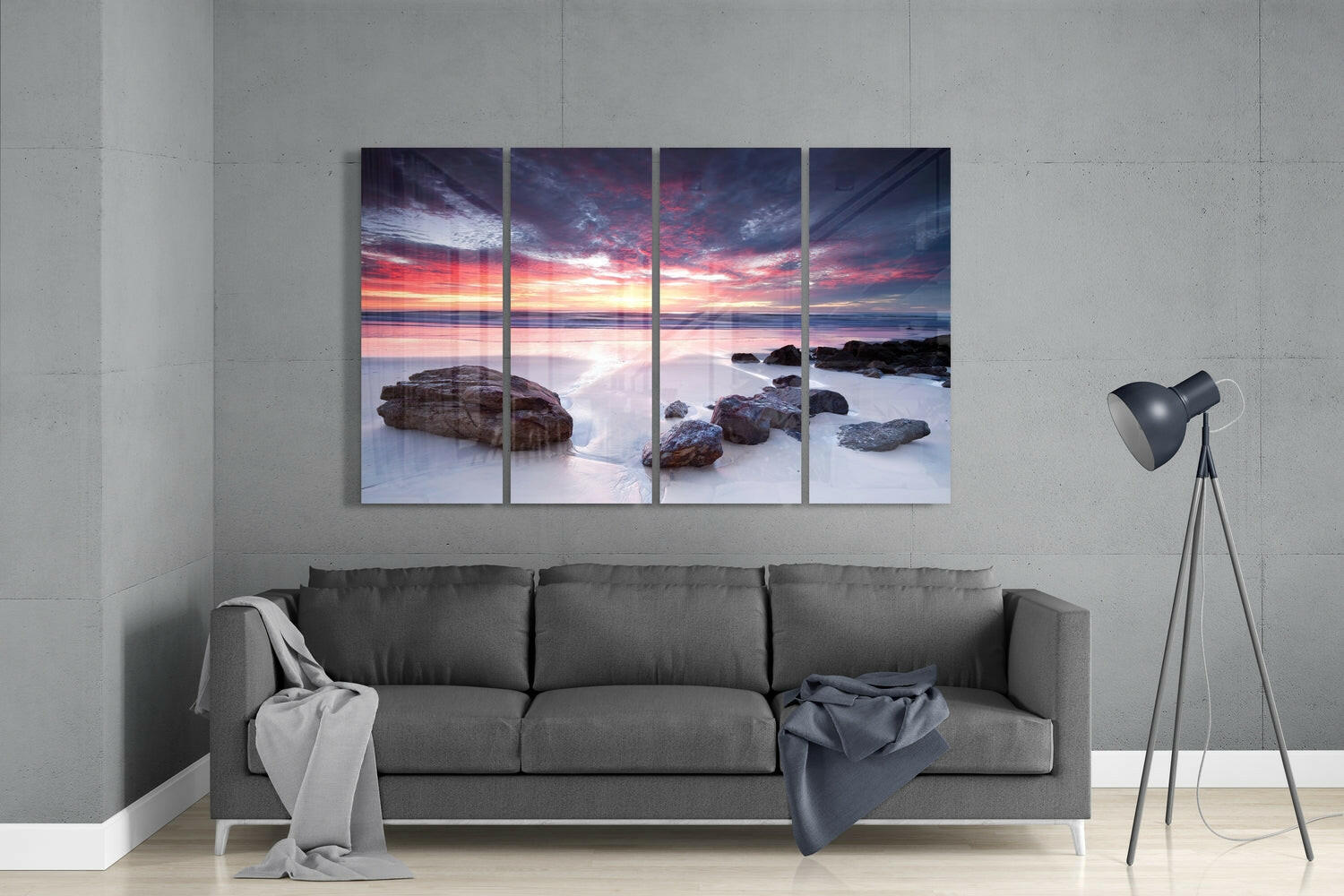 Four-panel glass wall art displaying a coastal sunset scene with pink and orange sky, rocky beach, and smooth water, mounted above a gray sofa in a modern living room.