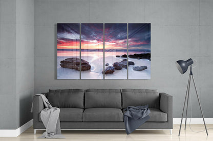 Four-panel glass wall art displaying a coastal sunset scene with pink and orange sky, rocky beach, and smooth water, mounted above a gray sofa in a modern living room.