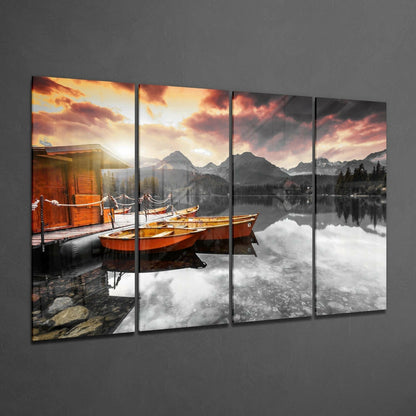 Four-panel glass wall art displaying orange boats moored at a wooden dock with mountains and sunset sky reflected in calm lake water.