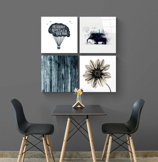 Four framed glass wall art prints displayed in a grid above a small black table with two black chairs, featuring a hot air balloon quote, elephant silhouette, blue wood texture, and sepia flower.