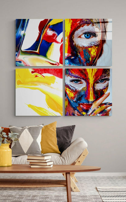 Four colorful glass panels featuring painted face designs in red, yellow, and blue mounted on a gray wall above a wooden bench with pillows and books.
